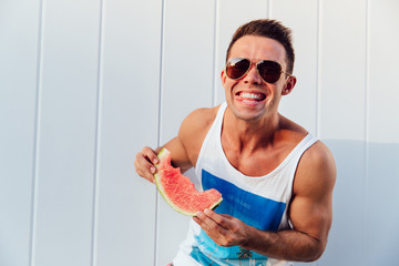 Wall Mural - Summer photo of smiling sportive man with watermelon, enjoying eating, looking at camera, wearing sunglasses, outdoors.