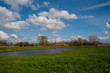 © heidepinkall - typische Landschaft der Wesermarsch mit grüner Weide, einem Siel und lebhaftem blauen Himmel mit weißen Wolken und im Hintergrund einem traditionellen Bauernhaus