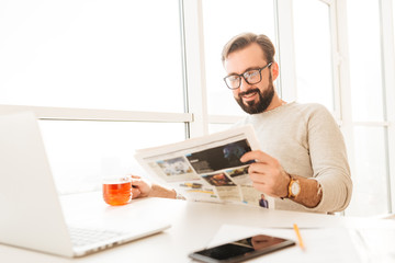 Sticker - Brunette bearded man wearing eyeglasses drinking tea from glass and reading newspaper, while resting at home