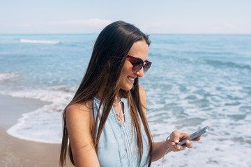 Portrait of smiling girl with long brown hair and sunglass on her face looks in her cell phone. Sea waves on background