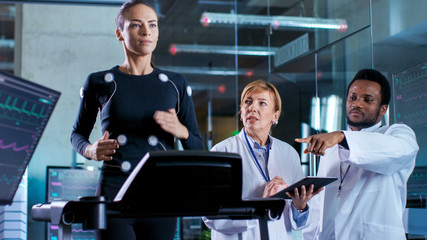 Wall Mural - beautiful woman athlete runs on a treadmill with electrodes attached to her body, while two scientis