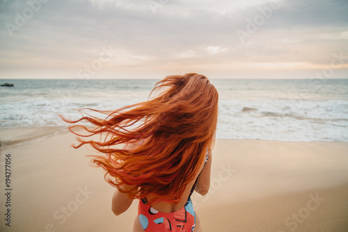 Fotografía young red-haired woman with flying hair on the ocean coast at sunset, rear view