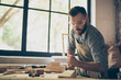 Confident professional busy hardworking bearded handsome serious wearing checkered casual shirt apron and protective goggles is aligning the surface of wooden plank with a plane near window
