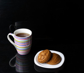 two oatmeal cookies lie on a white plate, next is a large striped mug with tea, on a black background