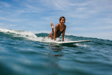 Beautiful young indonesian woman in bikini surfing wave in Bali on the background of blue sky, clouds and tropical beach