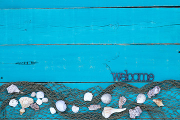 Wall Mural - Welcome sign hanging on antique rustic teal blue wood sign with seashells and fish netting border; wooden background with painted copy space