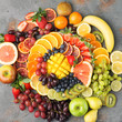 Colorful cut fruits platter in rainbow colors oranges grapes mango strawberries kiwis blueberries grapefruit on the grey concrete table arranged in circle, top view, selective focus