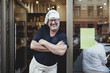 © Maskot - Smiling male baker standing with arms crossed at entrance of bakery