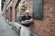© Maskot - Portrait of senior male baker leaning on brick wall outside bakery