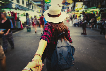 Young Asian woman with hat and backpack traveling in Khaosan Road among walking people, woman guiding man by the hand into outdoor market in Bangkok, Thailand.