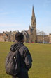 Turysta w ciemnym ubraniu, z plecakiem na plecach, tyłem, przygląda się wysokiej wieży zabytkowego kościoła w Edynburgu, Szkocja, zielony trawnik, zabudowa miejska w tle, błękitne niebo