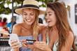 © WHstudio Leushin N - Cheerful best friends women meet together at cafeteria, happy to make online shopping with smart phone and plastic card, choose new clothes on internet website, happy to see something appropriate