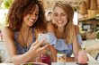 © WHstudio Leushin N - Happy young women watch funny video in internet on smart phone, sit together against cafe interior, eat tasty dessert and fresh cocktails. People, relationship and modern technology concept.