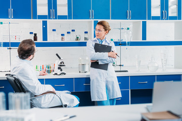 Wall Mural - partial view of female scientist in eyeglasses with notepad in hands looking at colleague at workplace in lab