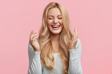 Wall Mural - Isolated shot of joyful blonde young cute woman laughs joyfully as hears funny anecdote from friend, has long light hair, poses against pink studio wall. Happiness and positive emotions concept