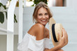 © WHstudio Leushin N - Horizontal shot of adorable smiling female wears fashionable white blouse, keeps straw summer hat in hands, spends free time at hotel as recreats in hot exotic country during unforgettable vacations