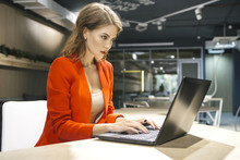 Beautiful Rich Caucasian Business Woman Sitting In Her Modern Office In Luxury Red Jacket. Neutral Interior With Dim Led Lighting. Woman Smiles Friendly, Working On A Laptop