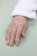 Stara zniszczona skóra dłoni bardzo starej kobiety w białym szlafroku na jasnym tle.