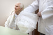 Pielęgniarz pokazuje pielucho majtki dla starych osób. Staruszka w białym szlafroku i koszuli nocnej siedzi na krześle na drugim planie.
