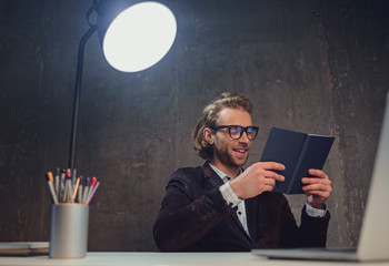 Portrait of happy bearded smart male reading book while situating at desk. Rest during job concept