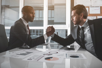 Who is stronger. Side view profile of two managers are sitting opposite each other with one elbow resting on table, clasp each other's hands, and trying to force each other's arm down onto the desk