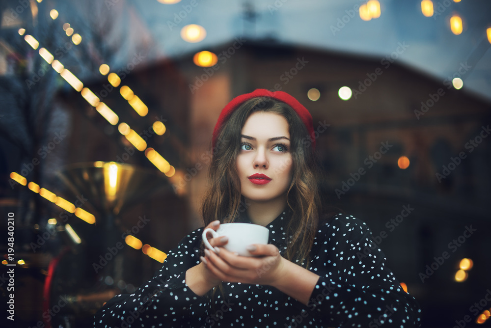 Young beautiful fashionable girl with red lips, wearing french style beret and polka dot blouse drinks coffee or tea in cafe. Model looking up. Copy, empty space for text - obrazy, fototapety, plakaty 