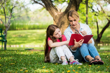 Wall Mural - Mother and daughter reading a book together in a park. Family, lifestyle and education concept