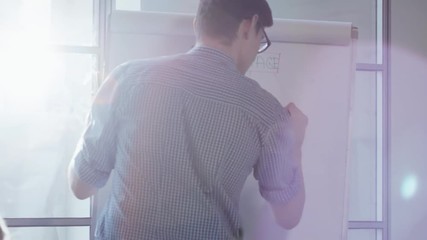 Young male teacher in glasses writing and drawing schemes on flip chart paper during coding computer class