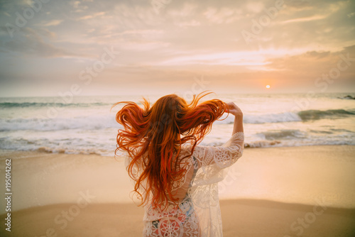 Fotomural young red-haired woman with flying hair on the ocean coast at sunset, rear view