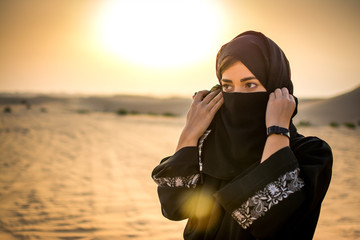 Wall Mural - Portrait of a young Arab woman wearing traditional black clothing in the desert during sunset.
