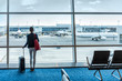 © Maridav - Traveler businesswoman waiting for delayed flight at airport lounge standing with luggage watching tarmac at airport window. Woman at boarding gate before departure. Travel lifestyle.