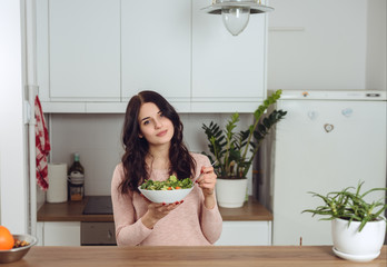 Smiling young woman in the kitchen near desk and eats vegetable salad. Healthy life concept