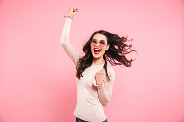 Wall Mural - Image of cheerful woman 20s in casual clenching fists with delight and smile, isolated over pink background