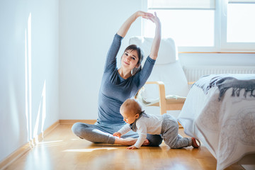 Wall Mural - Cute european mother doing yoga or pilates while her infant baby boy playing near at cozy simple home interior. Motherhood, healthy lifestyle and people concept.