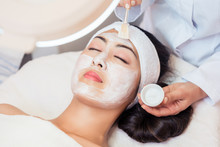 Close-up Of The Face Of A Young Woman Relaxing Under The Gentle Touch Of The Specialist, Applying On Her Cheeks White Facial Mask With Rejuvenating Effects