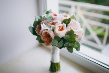 Close up A beautiful tender bridal bouquet in rose colors of pion-shaped roses and green twigs tied with a long silk white ribbon on a beige windowsill. Wedding accessories for bride