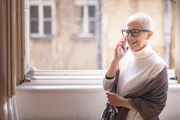 Beautiful senior lady talking on her cell phone, standing next to a big home window, enjoying her chat