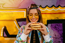 Attractive Young Woman, Smiling Cheerfully, Holds Tasty Burger In Two Hands. Dressed In Colorful Jacket And Cap, In Sunglasses. Outdoors, Near The Wall With Graffiti.