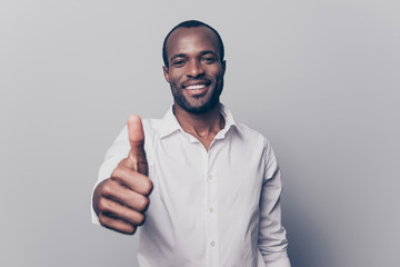 Portrait of cheerful, positive, handsome man with black skin, beaming smile in white shirt showing thumb up with finger to the camera isolated on grey background