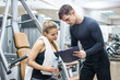 Young sporty woman and her personal trainer looking together at sports results on clipboard in gym.
