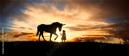 Valokuva  a sweet little girl offers fresh grass the Unicorn in a meadow at sunset