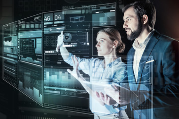 Creative colleagues. Smart qualified young programmer carefully touching the transparent screen while her friendly reliable colleague standing near and looking at it