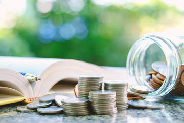 Pile of money coins in and outside the glass jar with the book on blurred natural green background