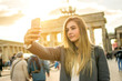 Beautiful blond young woman taking selfie in front of Brandenburg Gate in the city of Berlin, Germany.