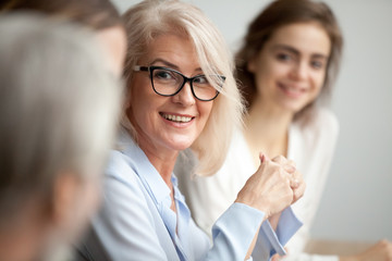 Smiling aged businesswoman in glasses looking at colleague at team meeting, happy attentive female team leader listening to new project idea, coach mentor teacher excited by interesting discussion