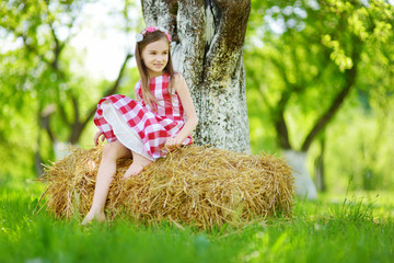 Wall Mural - Adorable little girl sitting on a haystack in apple tree garden on warm and sunny summer day