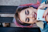 Fototapeta  - Close-up portrait of a smiling girl with dyed hair using phone while lying on a bench. Top view.