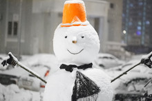 Snowman On A Blurred Background In Gray With Blue Lights Of The Cityscape. Closeup Of Smiling Snowman With Orange Hat, Scarf And Carrot Nose, Outdoors In Snowfall. New Year Snow Concept Happy Holiday