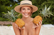 © WHstudio Leushin N - Outdoor shot of beautiful woman wears straw hat, poses nude on tropical beach with two exotic pineapples, smiles joyfully, enjoys summer vacation abroad. Positive female vegeterian eats healthy food