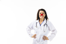 Portrait Of A Smiling Female Doctor With A Red Clown Nose And Both Arms On Her Waist, Isolated On White Background